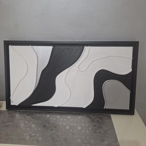 May include: Abstract wall art in a black frame. The artwork features a layered design with black, white, and gray organic shapes. The piece has a modern aesthetic, suitable for home or office decor.
