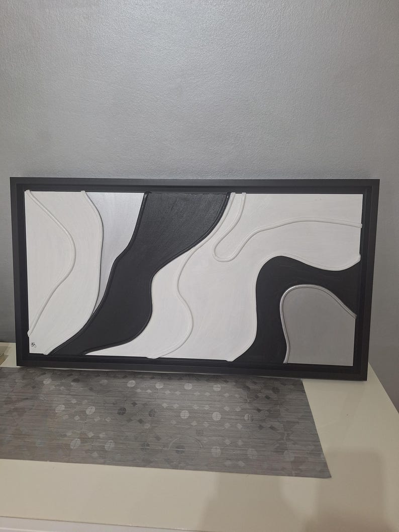 May include: Abstract wall art in a black frame. The artwork features a layered design with black, white, and gray organic shapes. The piece has a modern aesthetic, suitable for home or office decor.