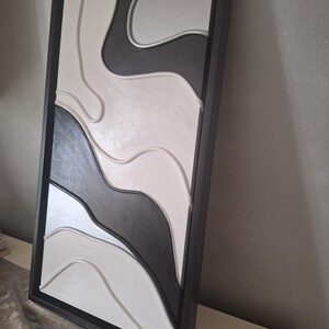 May include: Abstract wall art in a black frame. The artwork features a layered design with flowing, organic shapes in shades of black, white, and gray. The piece has a modern, minimalist aesthetic, suitable for home decor.