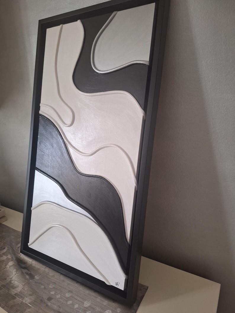 May include: Abstract wall art in a black frame. The artwork features a layered design with flowing, organic shapes in shades of black, white, and gray. The piece has a modern, minimalist aesthetic, suitable for home decor.