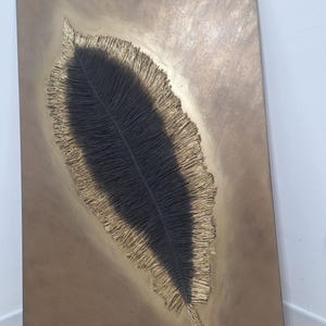 May include: A rectangular wall art piece featuring a large, textured feather design. The feather is black with a gold-colored, fringed border, set against a bronze-toned background. The artwork has a modern, abstract aesthetic.