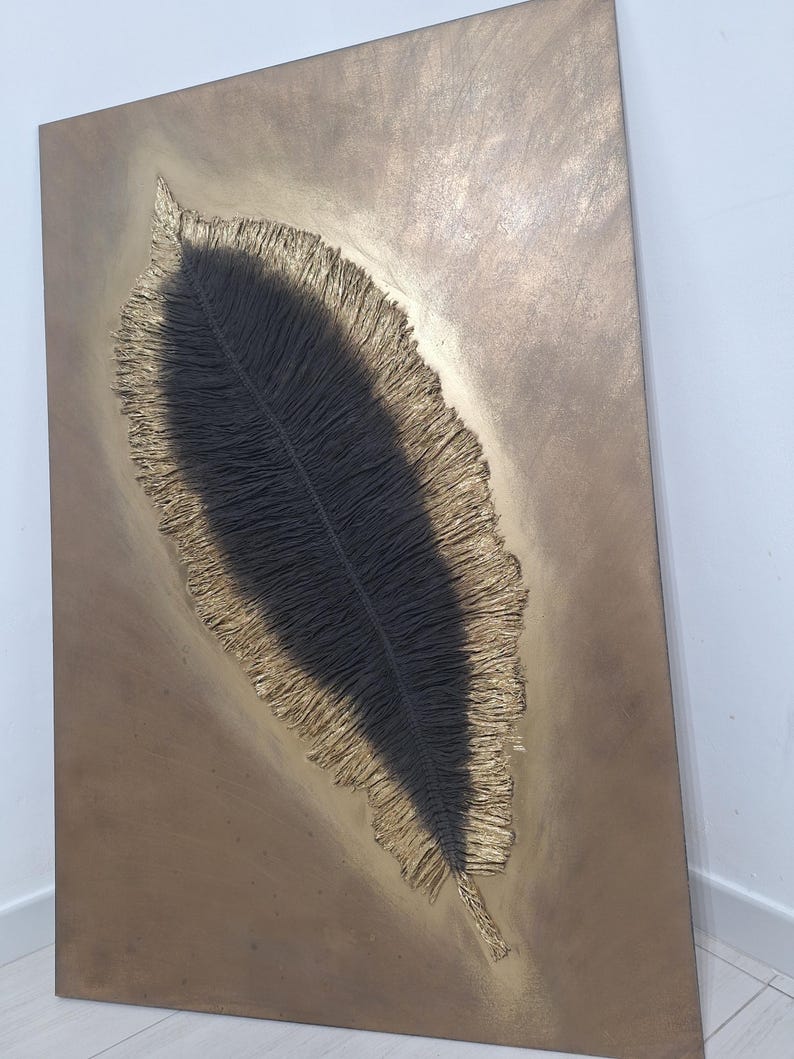 May include: A rectangular wall art piece featuring a large, textured feather design. The feather is black with a gold-colored, fringed border, set against a bronze-toned background. The artwork has a modern, abstract aesthetic.