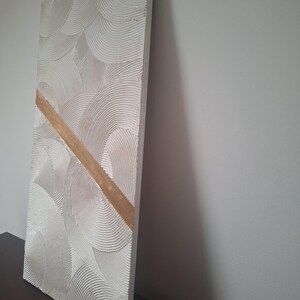 May include: A rectangular, textured, off-white wall art piece with a repeating semi-circular pattern. A diagonal gold-colored stripe bisects the artwork. The piece is displayed against a neutral wall, showcasing its three-dimensional texture and design.