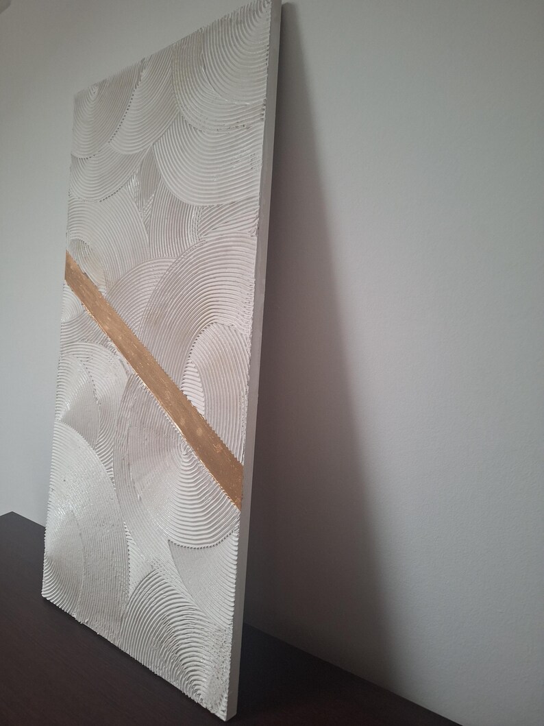 May include: A rectangular, textured, off-white wall art piece with a repeating semi-circular pattern. A diagonal gold-colored stripe bisects the artwork. The piece is displayed against a neutral wall, showcasing its three-dimensional texture and design.