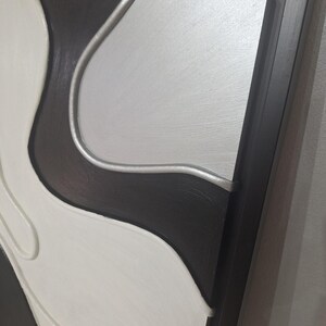 May include: Abstract wall art in a black, white, and silver color scheme. The artwork features a layered design with curved shapes and a dark wooden frame. The piece has a modern, minimalist aesthetic.