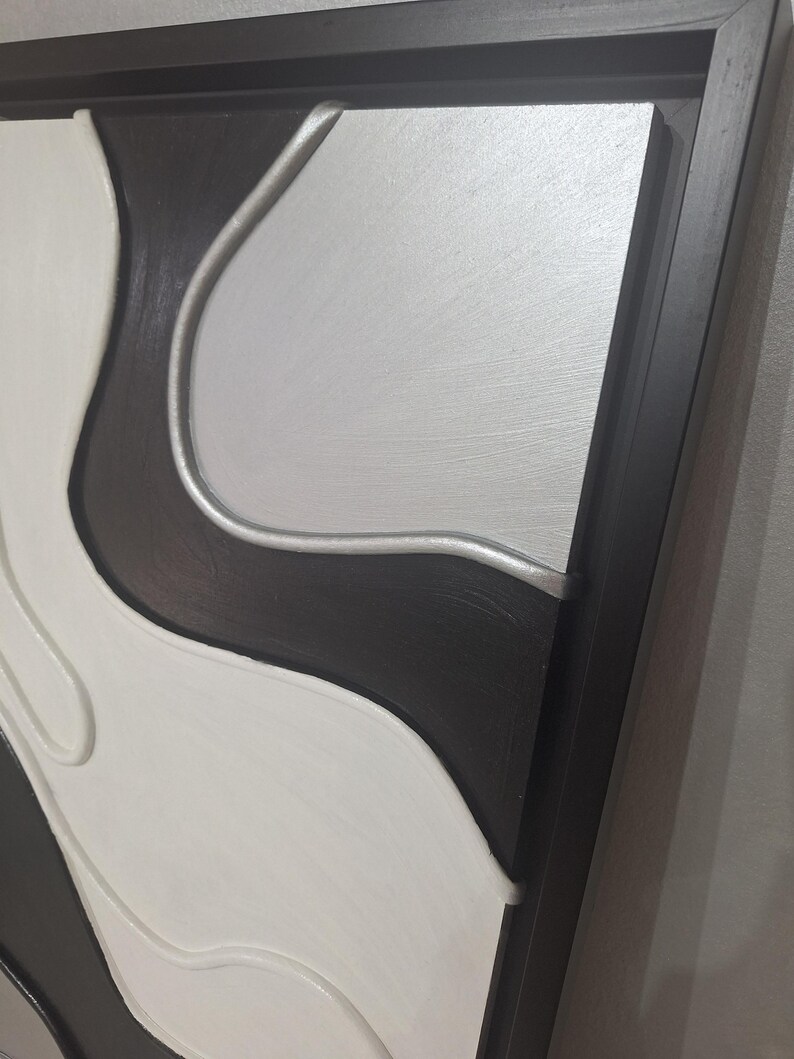 May include: Abstract wall art in a black, white, and silver color scheme. The artwork features a layered design with curved shapes and a dark wooden frame. The piece has a modern, minimalist aesthetic.