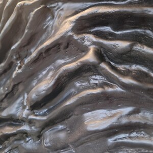 May include: Close-up of a textured, dark gray sculpture with a glossy finish. The surface has a rippled, organic appearance, with highlights and shadows creating depth. The sculpture appears to be made of a solid material.