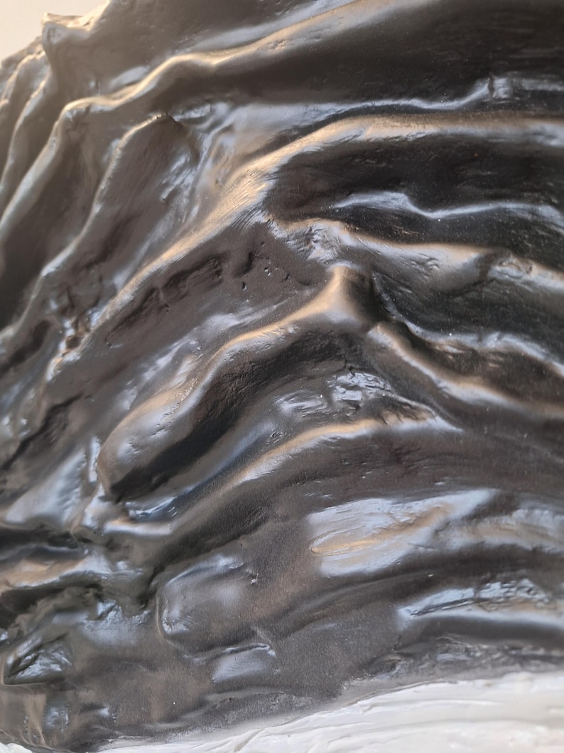 May include: Close-up of a textured, dark gray sculpture with a glossy finish. The surface has a rippled, organic appearance, with highlights and shadows creating depth. The sculpture appears to be made of a solid material.