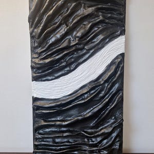 May include: Abstract textured wall art. The rectangular piece features a black, rippled surface with a white, textured stripe running diagonally across it. The artwork has a modern, minimalist aesthetic.