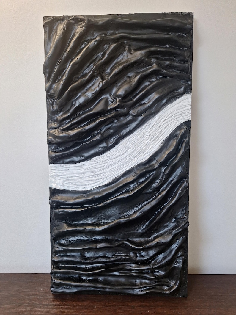 May include: Abstract textured wall art. The rectangular piece features a black, rippled surface with a white, textured stripe running diagonally across it. The artwork has a modern, minimalist aesthetic.