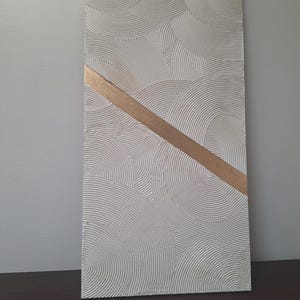 May include: Textured white rectangular wall art with a gold diagonal stripe. The white surface features a repeating pattern of curved lines, creating a layered, circular design. The gold stripe adds a contrasting element to the neutral color palette.