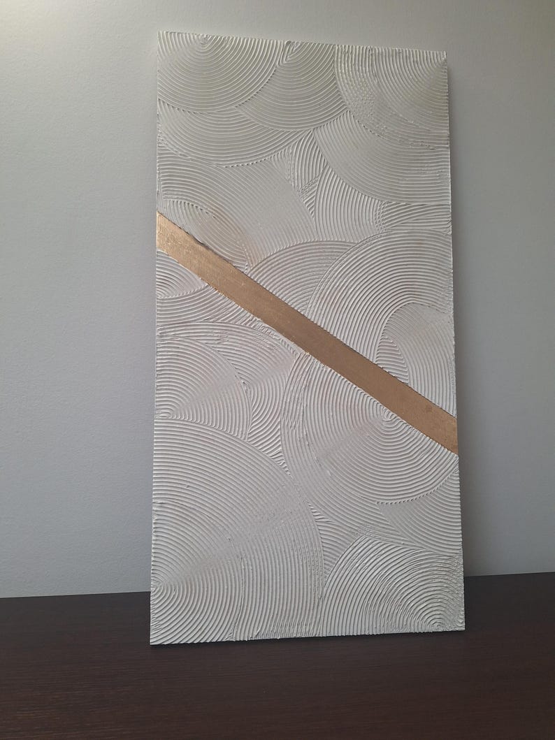 May include: Textured white rectangular wall art with a gold diagonal stripe. The white surface features a repeating pattern of curved lines, creating a layered, circular design. The gold stripe adds a contrasting element to the neutral color palette.