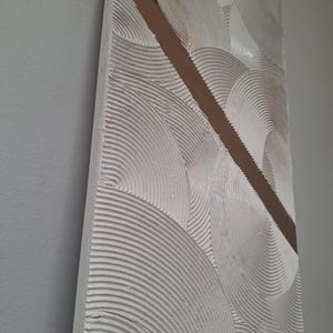 May include: Textured white wall art with a diagonal brown stripe. The artwork features a repeating pattern of semi-circular, ridged designs. The piece is mounted on a rectangular wooden frame, creating a modern, abstract aesthetic.