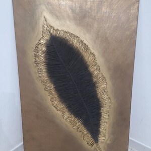 May include: A rectangular, bronze-toned artwork featuring a textured, stylized feather. The feather's central vein is dark, surrounded by a gold-colored, fringed edge. The background has a mottled, metallic finish, creating a striking contrast.