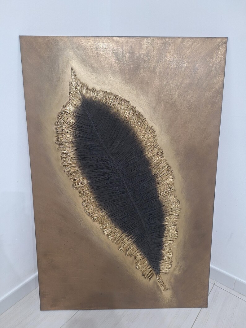 May include: A rectangular, bronze-toned artwork featuring a textured, stylized feather. The feather's central vein is dark, surrounded by a gold-colored, fringed edge. The background has a mottled, metallic finish, creating a striking contrast.