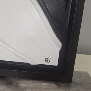 May include: Abstract artwork in a black frame. The piece features a textured, layered design with contrasting black and white sections. A small, stylized signature is visible in the lower corner. The artwork is displayed on a light-colored surface.