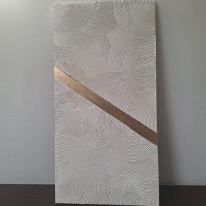 May include: A rectangular, textured, white wall art piece with a repeating circular pattern. A diagonal band of gold-colored material crosses the artwork. The piece is displayed against a neutral background, showcasing its minimalist design.