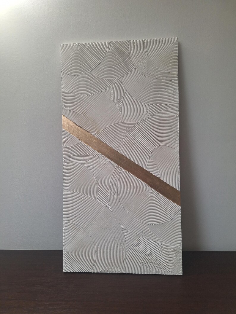 May include: A rectangular, textured, white wall art piece with a repeating circular pattern. A diagonal band of gold-colored material crosses the artwork. The piece is displayed against a neutral background, showcasing its minimalist design.
