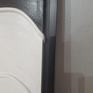 May include: Close-up of a section of a framed artwork. The artwork features a white, curved design against a textured black background. A black frame surrounds the artwork, with a section of a white wall visible.