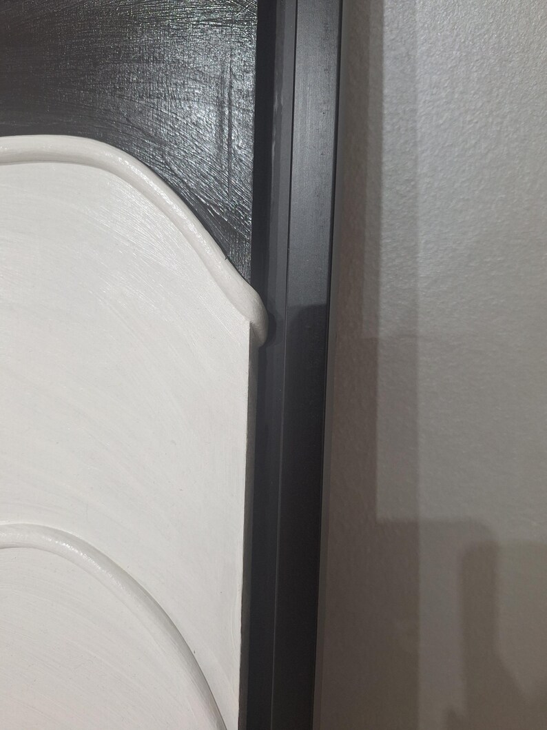 May include: Close-up of a section of a framed artwork. The artwork features a white, curved design against a textured black background. A black frame surrounds the artwork, with a section of a white wall visible.