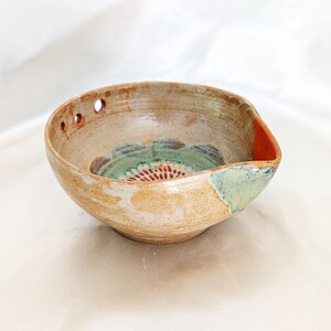 Peut inclure: Un bol en céramique rond avec un extérieur beige texturé et un bec verseur. L'intérieur présente un motif floral vert et orange à la base. Deux petits trous circulaires sont situés sur le bord supérieur près de la zone du bec verseur.