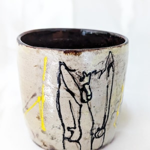 Peut inclure: Une tasse en céramique avec un émail blanc cassé moucheté, dotée d'un intérieur et d'un bord brun foncé. L'extérieur est orné de dessins au trait noir représentant une silhouette et de traits de peinture jaune vif verticaux.