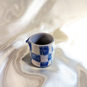 May include: A small, ceramic pitcher with a blue and white checkered pattern. The pitcher has a spout and a light blue interior. The handmade pottery sits on a white, silky surface.