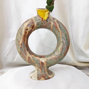 Peut inclure: Un vase en céramique circulaire avec une ouverture centrale, présentant un émail marbré dans des tons de vert sauge, brun et crème. Le bord supérieur est accentué par un émail jaune vif, et une tige feuillue verte est placée dans l'ouverture.