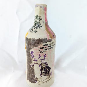 May include: Hand-painted ceramic bottle with a unique design. The bottle has a textured, off-white base with pink, brown, yellow, and black accents. The bottle features a drawing of a person and a dog, with the text "Well maybe just a nice shade of pink."