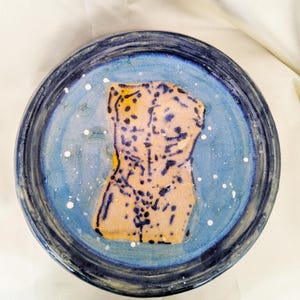 May include: A hand-painted ceramic plate with a dark blue rim and a light blue centre. The centre features a yellow and black torso design with white speckles. The plate is round and appears handmade.