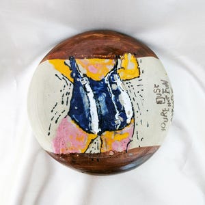 Peut inclure: Une assiette en céramique peinte à la main avec un bord marron et un centre blanc. L'assiette présente une illustration fantaisiste d'une personne en maillot de bain bleu et blanc, avec des accents roses et jaunes. Le texte sur l'assiette dit "You're just not fun anymore."