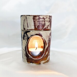 May include: A cylindrical ceramic oil warmer featuring a mottled cream and dark brown glaze. The structure consists of a base with a large oval cutout containing a lit tea light candle and a removable top dish for wax or oil warming.
