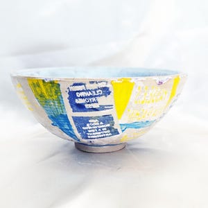 Peut inclure: Un bol en céramique avec un intérieur bleu clair et un extérieur texturé présentant un collage d'éléments de texte imprimés en jaune, bleu et blanc. Le bol possède un large bord circulaire et une petite base surélevée, affichant un aspect superposé.