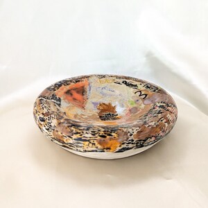 May include: An oval ceramic bowl with a white base. The interior is a collage of colours including orange, brown, black, and beige. The rim has a speckled pattern. The number "3" is visible on the interior. The bowl is handmade.