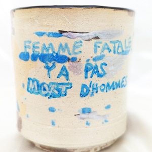 Puede incluir: Una taza de cerámica cilíndrica con un acabado beige moteado. El exterior presenta un texto azul pintado a mano que dice "FEMME FATALE Y A PAS MORT D'HOMMES" junto con trazos y manchas de pintura azul y plateada por la superficie.