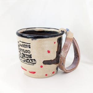 Può includere: Una tazza in ceramica fatta a mano con smalto color crema, caratterizzata da una scritta nera dipinta a mano che recita LOOKING FORWARD TO BEING ATTACKED. La tazza presenta un bordo marrone scuro, puntini rossi sparsi, una striscia astratta marrone scuro e un manico angolare testurizzato.