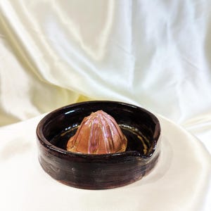 May include: A handmade ceramic citrus juicer. The juicer features a dark brown bowl with a spout and a central reamer in shades of brown and pink. The juicer is set against a white background.