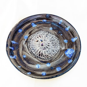 May include: A round ceramic plate featuring a dark brown and black glazed surface with a textured, light-colored circular center pattern. The plate is decorated with scattered, raised blue spots of varying sizes across the dark outer rim.