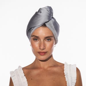 Pode incluir: Uma toalha de cabelo cinza enrolada na cabeça. A toalha é feita de um material macio e sedoso com uma borda de tecido felpudo. Projetada para absorver água e reduzir o frizz. A modelo está vestindo um top branco com babados.