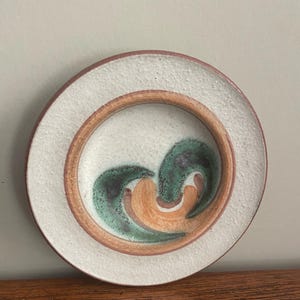Op de afbeelding: Een ronde keramische plaat met een getextureerd crèmekleurig oppervlak en een bruine rand. Het midden heeft een ontwerp van groene en oranje wervelingen. De plaat is waarschijnlijk handgemaakt.