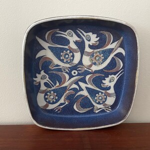 Op de afbeelding: Vierkante keramische schaal met een diepblauwe achtergrond en een witte rand. De schaal heeft een herhalend patroon van gestileerde witte vogels met bruine accenten en bloemendetails, wat een decoratief ontwerp creëert.