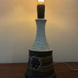 Op de afbeelding: Decoratieve tafellamp met een getextureerd, gebroken wit bovenstuk en een bruine, bloemversierde basis. De lamp heeft een vintage-stijl gloeilamp en een zwarte fitting. De lamp is aangesloten en brandt, waardoor een warme gloed ontstaat.
