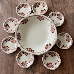 Op de afbeelding: Een vintage crèmekleurige servies set met bloemmotieven. De set bevat een grote serveerschaal en acht kleinere borden. Elk stuk heeft een bloemmotief in roze, blauwe, gele en rode tinten, met een gouden rand. De set is geplaatst op een houten oppervlak.