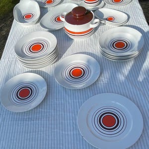 Peut inclure: Un service de table vintage en céramique blanche au design rétro. L'ensemble comprend des assiettes, des bols, un plat de service, une cocotte couverte et une tasse et une soucoupe. Chaque pièce présente un cercle orange central entouré d'anneaux concentriques noirs et marron.