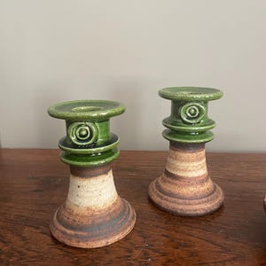 May include: Two ceramic candle holders. The candle holders have a green top with a spiral design, a tan and brown body, and a flared base. The candle holders are on a wooden surface.