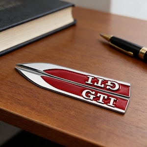 Puede incluir: Dos emblemas de coche GTI rojos y plateados. Los emblemas son alargados con un extremo puntiagudo plateado y un centro rojo. La palabra "GTI" se muestra en letras mayúsculas blancas en cada emblema. Los emblemas están sobre una superficie de madera.