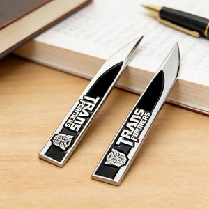 May include: Two silver and black Transformers logo letter openers. Each opener has a black centre with the Transformers logo and text. The openers are on a wooden surface next to a book and pen.