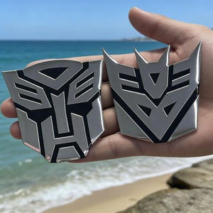 May include: Two silver emblems with black detailing, featuring geometric designs. The emblems are held up against a beach and ocean backdrop. The emblems are likely related to the Transformers franchise.