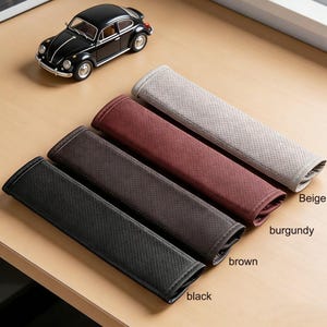 May include: Four car seat belt covers in black, brown, burgundy, and beige. Each cover is rectangular with a perforated design and a contrasting border. A black toy car is in the background.