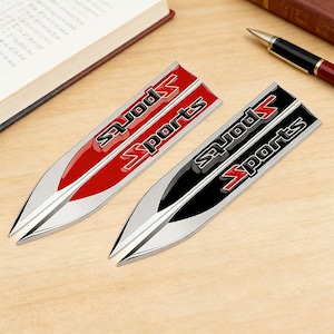 Pode incluir: Dois emblemas metálicos para carros com base prateada e design pontiagudo. Um é vermelho com texto "Sports" preto, o outro é preto com texto "Sports" vermelho. A palavra "Sports" está em uma fonte estilizada.